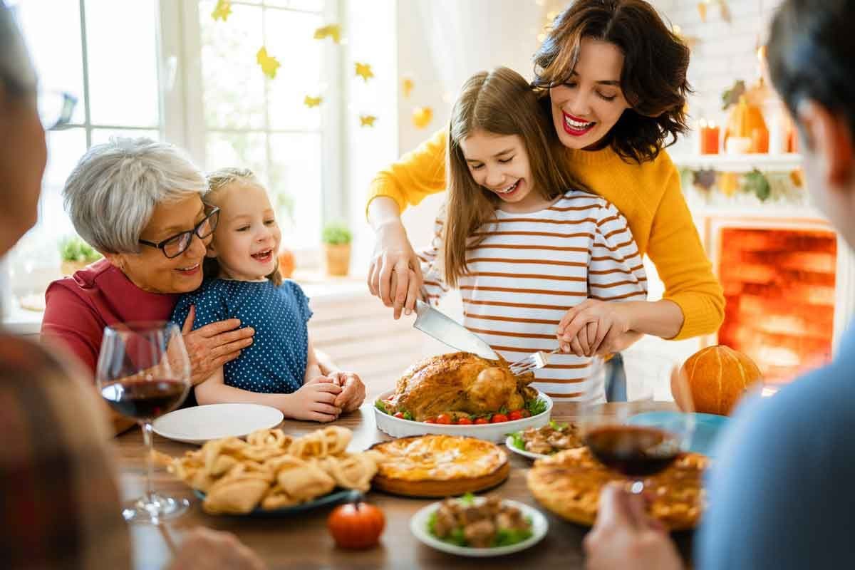 A family is gathered around the table for Thanksgiving, with a mother helping a daughter carve the turkey.