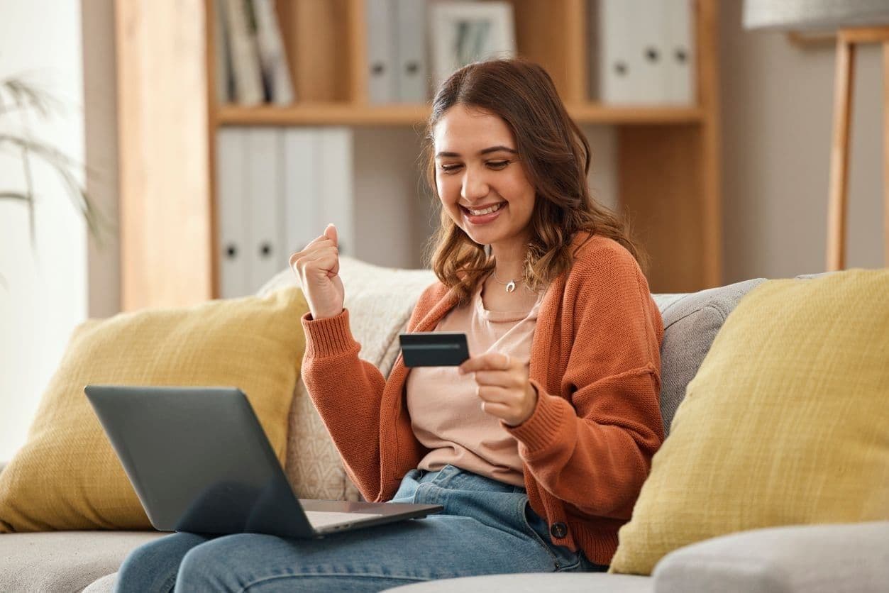 A woman sits on her sofa with a laptop in her lap and looks at her credit card.