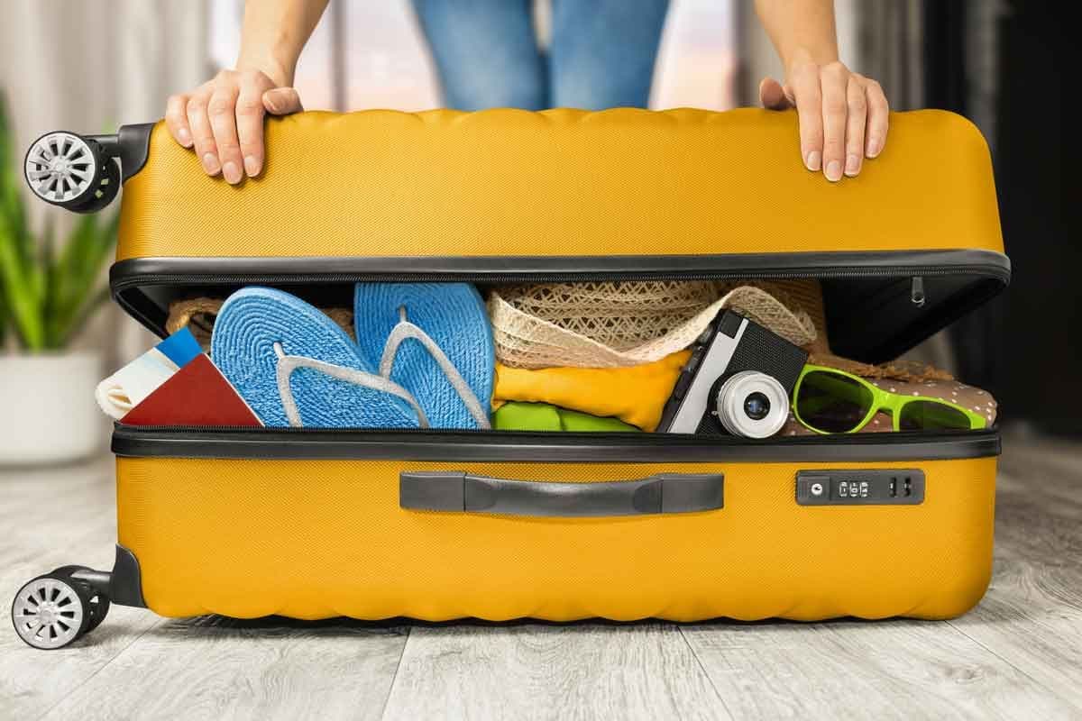 A woman's hands push down on a fully packed yellow suitcase with flip flops and other beach vacation gear.