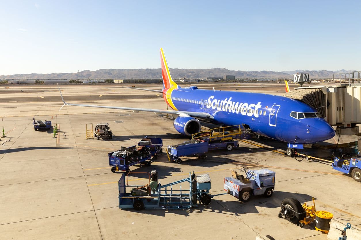 Southwest Airlines Boeing 737 parked at an airport gate, with ground crew equipment and luggage carts on the tarmac and mountains in the background.