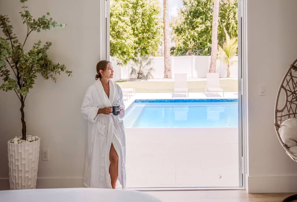 Woman in a white robe holding a coffee mug, looking out toward a pool from a bright hotel room doorway.