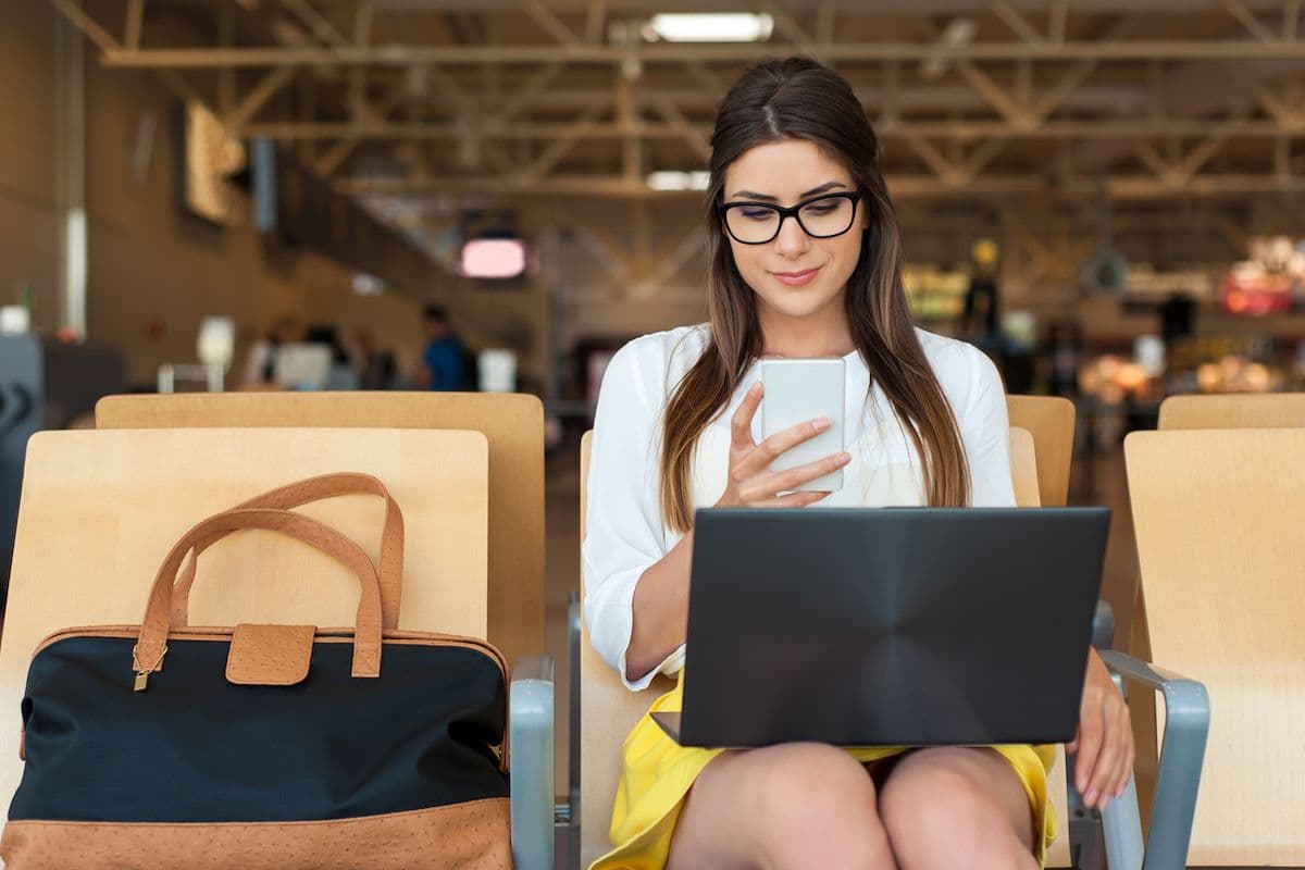 A woman sits in an airport terminal with a carry-on bag beside her, looking at her smartphone while working on a laptop at her seat.