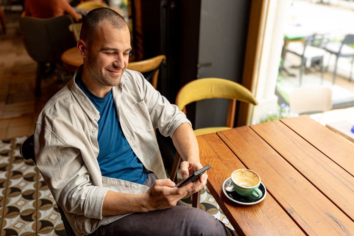 A man relaxes in a chair at a café, looking at his phone with a cappuccino on the wooden table next to him.