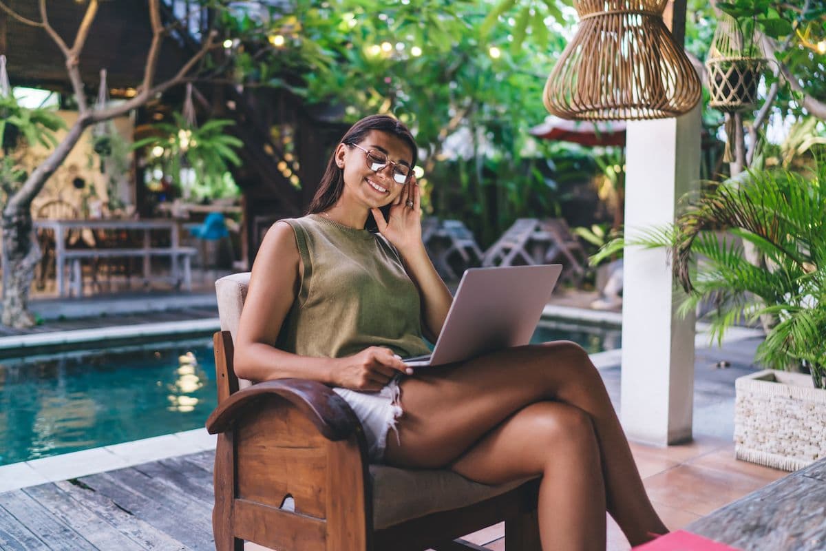 Smiling woman in sunglasses using a laptop while seated in a wooden chair beside a resort pool surrounded by tropical greenery
