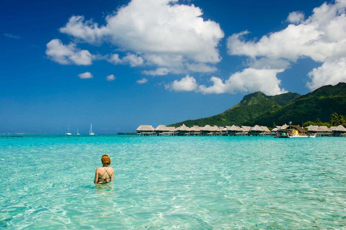 A woman stands in the ocean near her overwater bungalow.