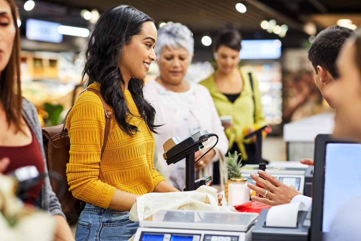 Smiling woman in a yellow sweater checks out at a busy grocery store register