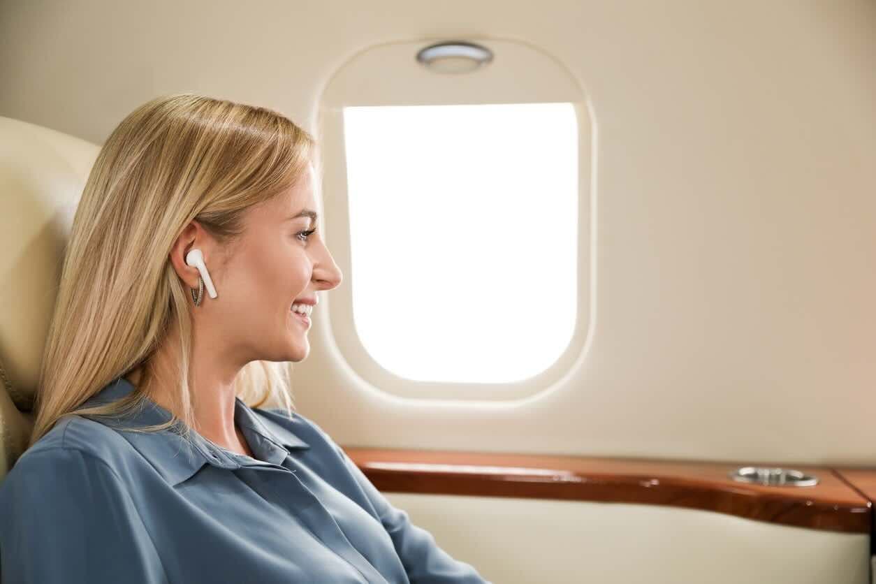 A woman in a window seat on an airplane wears ear bud headphones.