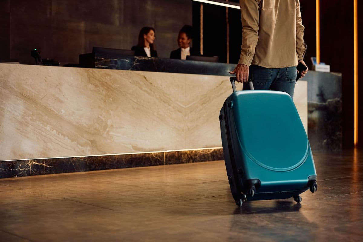 A hotel guest arrives at an upscale hotel lobby, rolling a teal suitcase toward the front desk