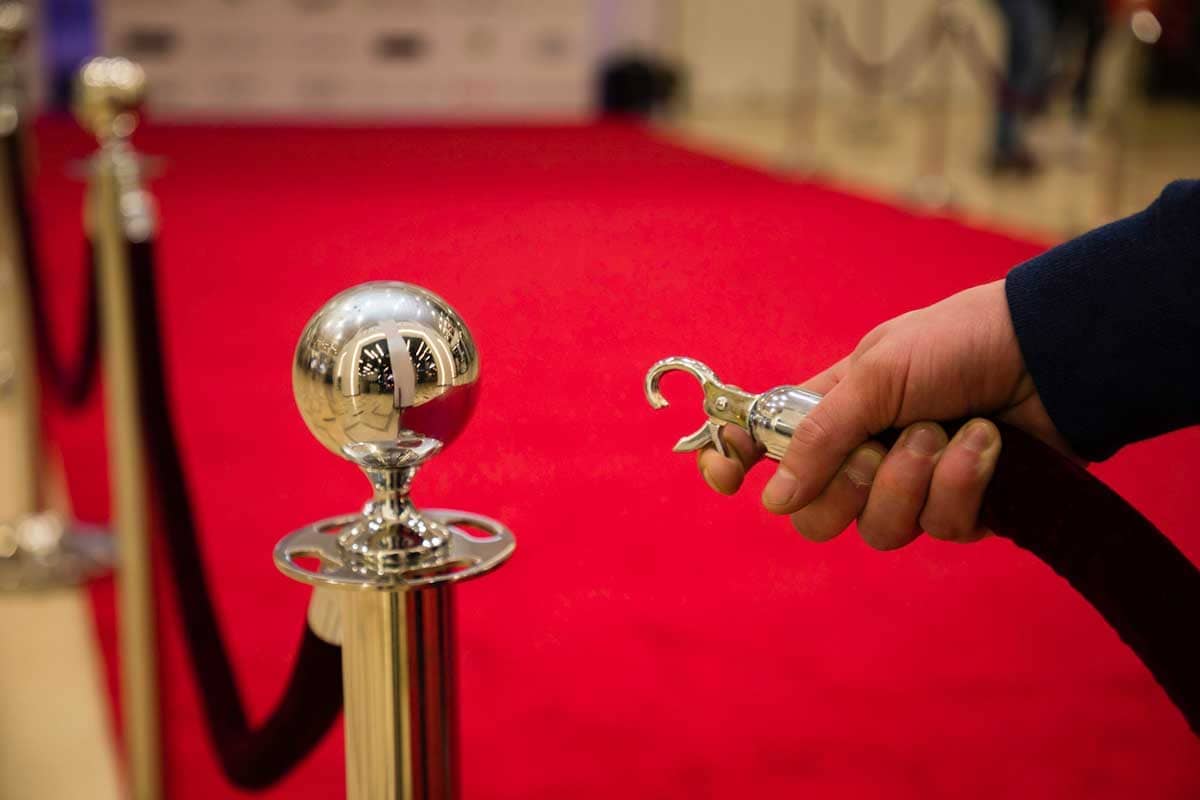 A hand unlocks a velvet rope in front of a red carpet.