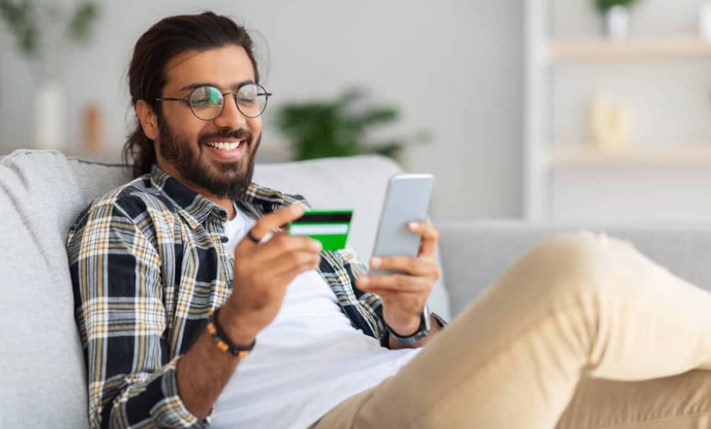 Smiling man on a couch holding a credit card and smartphone