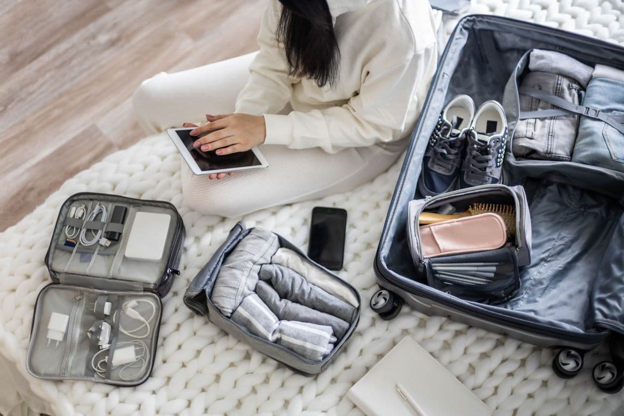 A woman sits on a bed next to an open suitcase, holding a tablet while organizing travel accessories including a tech organizer, packing cubes, and a toiletry bag.