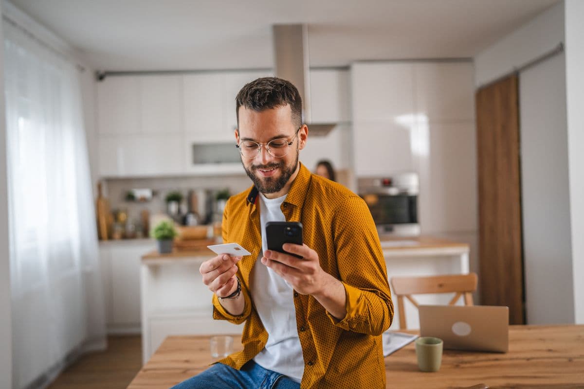 A man leans against his kitchen table while looking at his phone and holding a credit card to make a purchase.