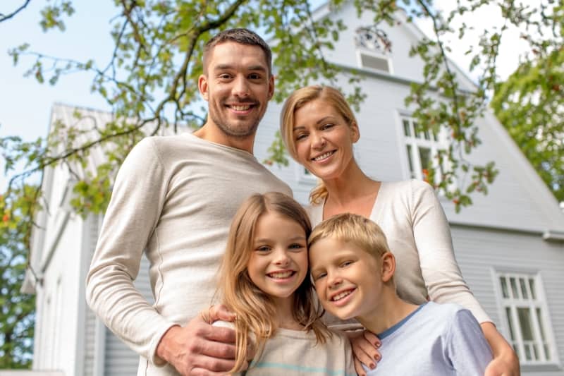 A Caucasian nuclear family smiles and stands in front of a suburban house