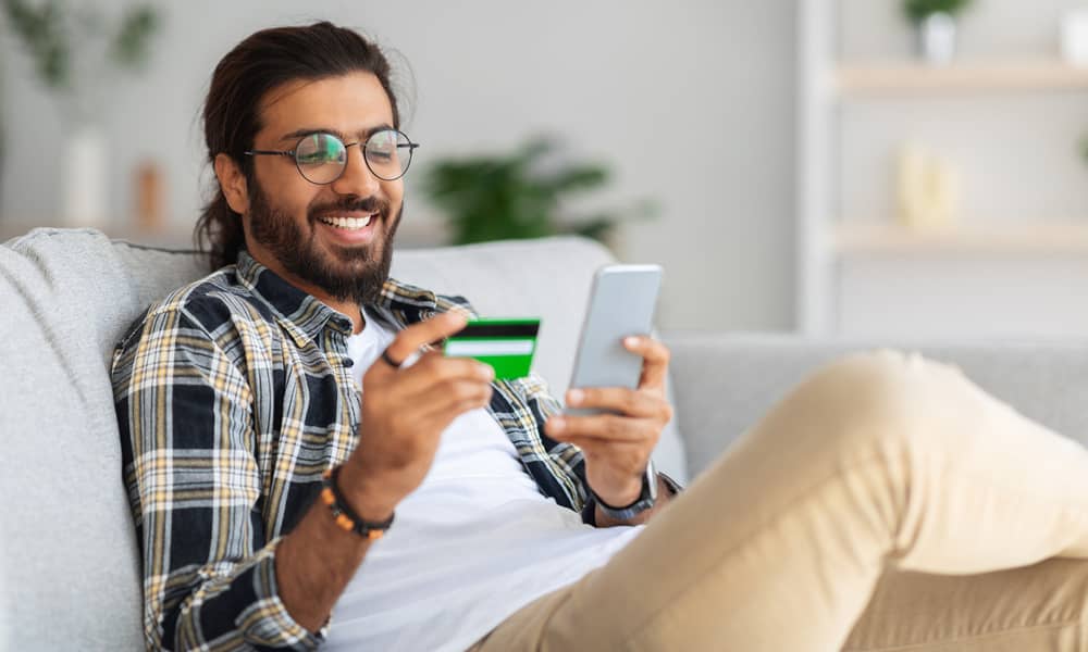 Smiling man relaxing on a couch, holding a credit card in one hand and a smartphone in the other.