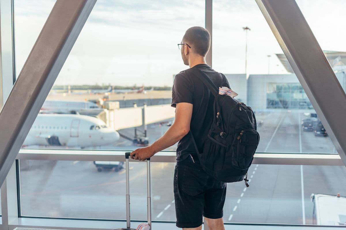 A man with a rolling suitcase and backpack stands at an airport gate window, looking out at two parked airplanes on the tarmac.