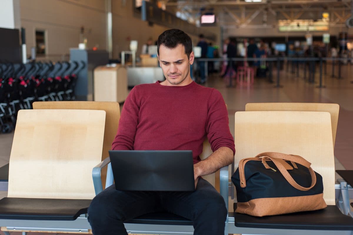 A traveler works on a laptop at an airport gate, with a travel bag on the seat beside him.