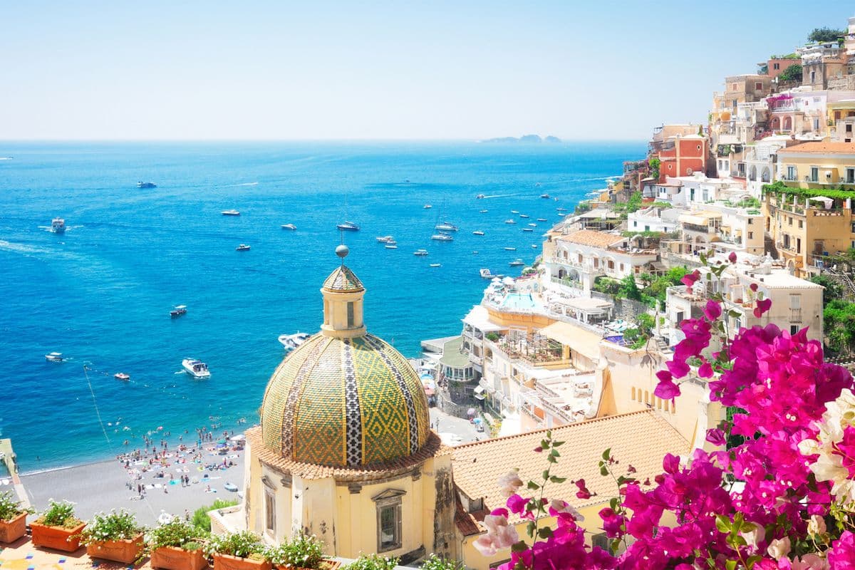 Aerial view of Positano, Italy, with a mosaic church dome, colorful cliffside buildings, pink bougainvillea, and the Mediterranean Sea