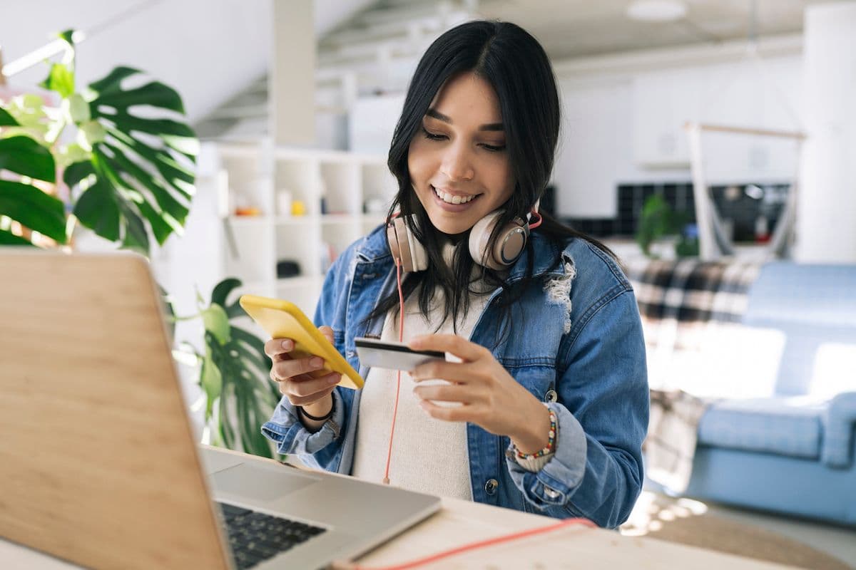 Smiling woman in a denim jacket sits at a desk holding a credit card and a phone, with a laptop open in front of her.