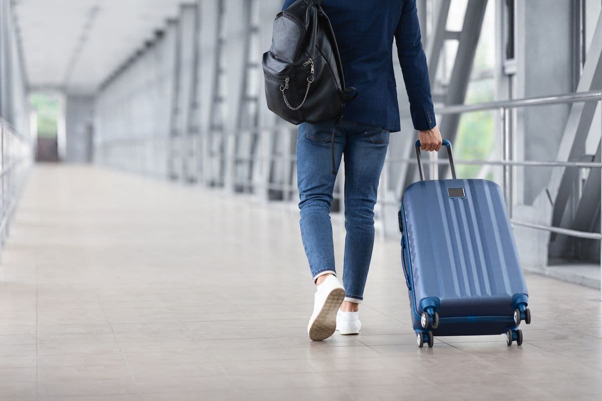 Rear view of a traveler in a blazer and jeans pulling a carry-on suitcase through an airport terminal