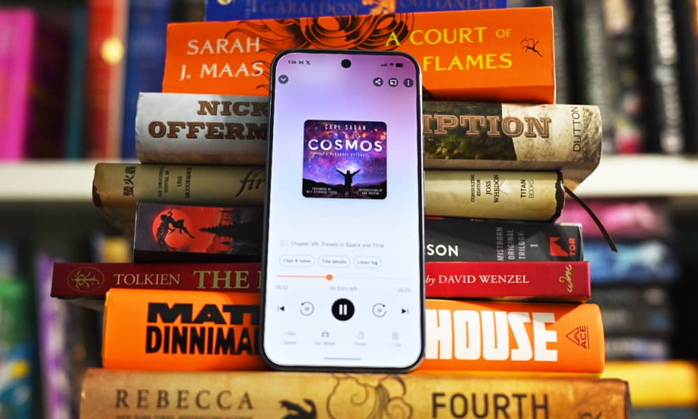 A smartphone propped upright against a tall stack of colorful hardcover books, its screen showing an the Audible app open mid-listen.
