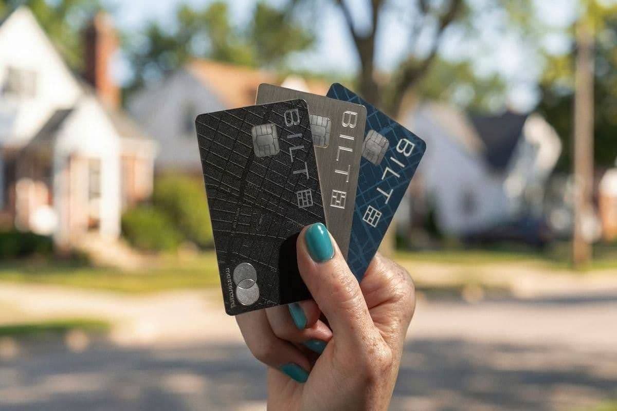 A hand holding three Bilt credit cards fanned out in front of a suburban neighborhood with houses and trees softly blurred in the background.