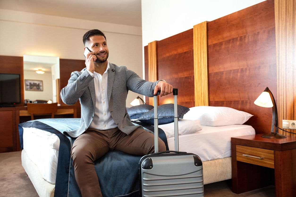 A man with a suitcase sits on a hotel bed talking on a cell phone
