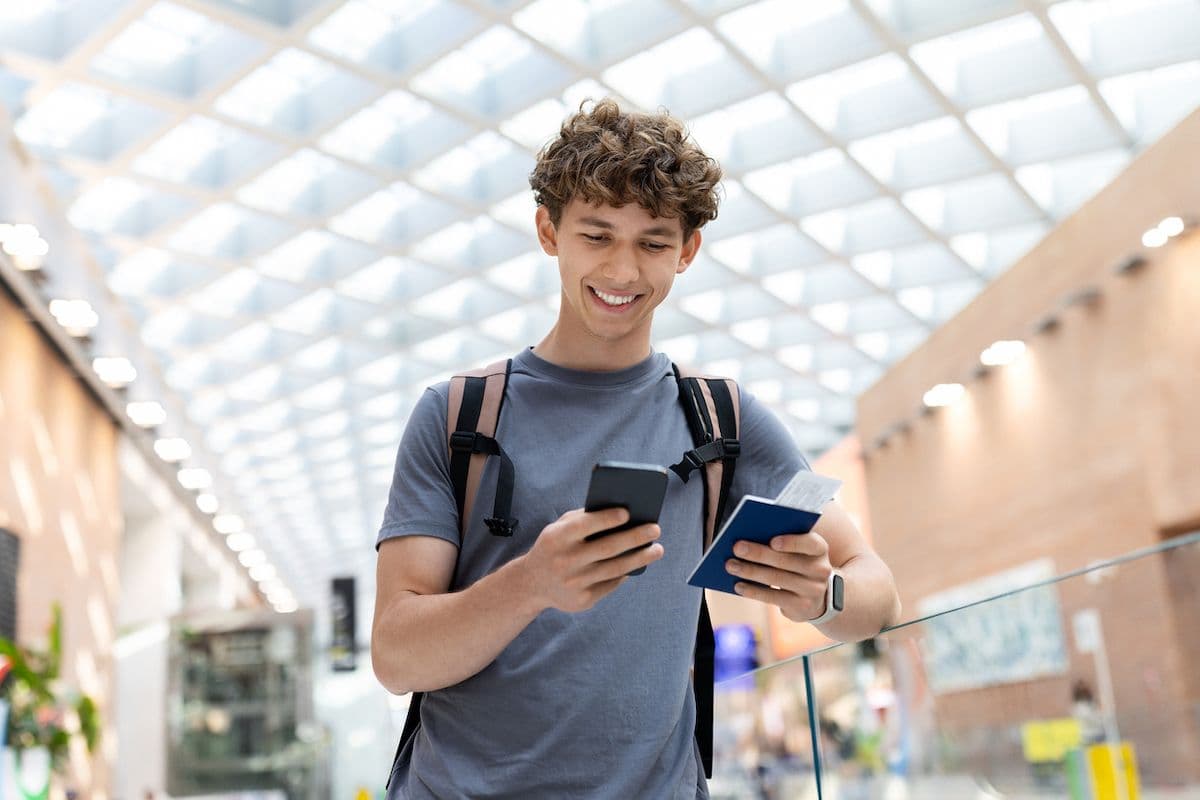 Smiling young traveler with a backpack checking his phone and holding a passport in an airport terminal