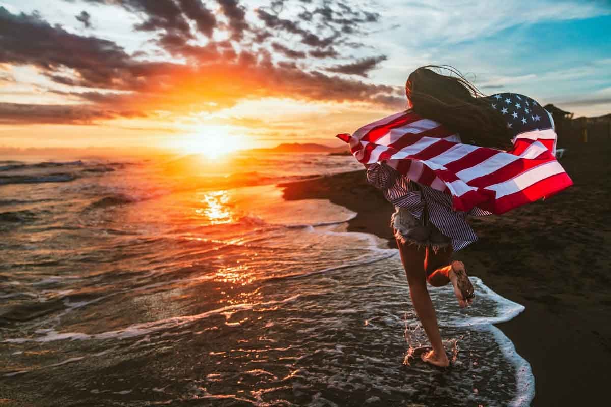 A young woman with the American flag held in her outstretched hands running beside the ocean.