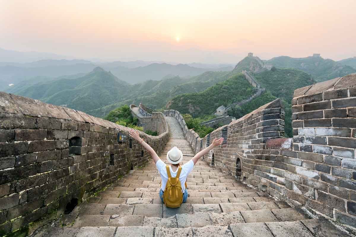 A tourist sits on the Great Wall of China with their hands raised.