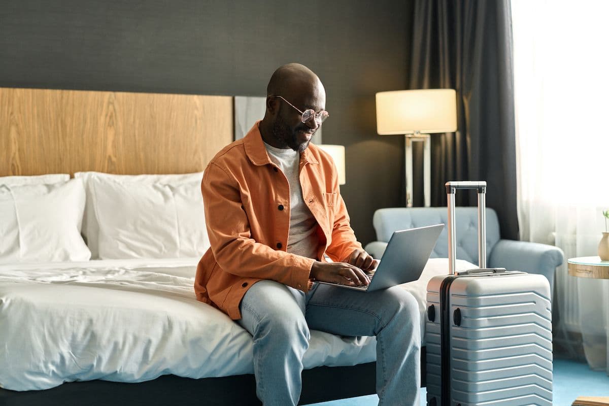 A man sits on the edge of a hotel bed while working on his laptop, with his suitcase next to him.