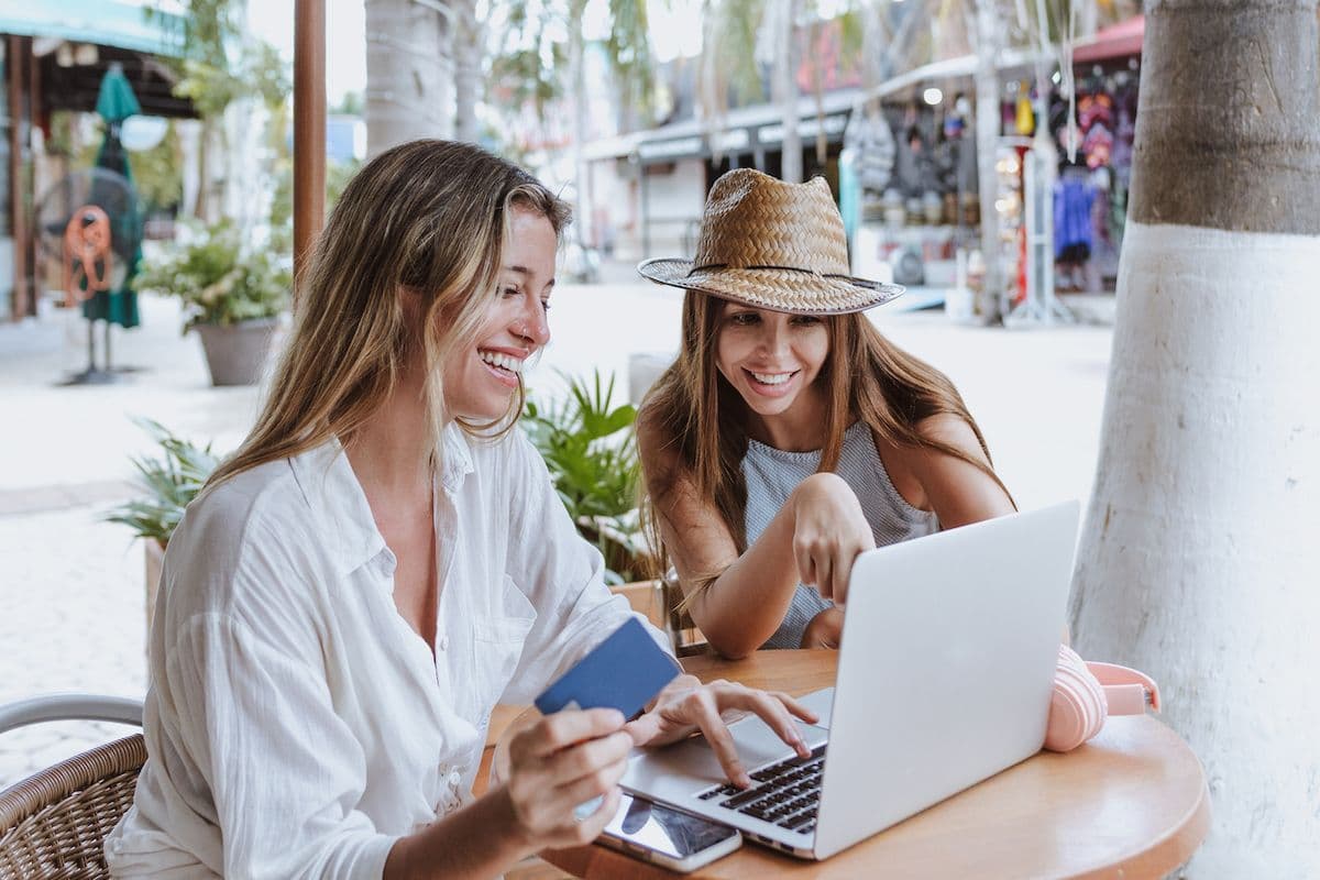 Smiling woman in white blouse holds a credit card while using a laptop at an outdoor café table, with a friend leaning in to look at the screen.