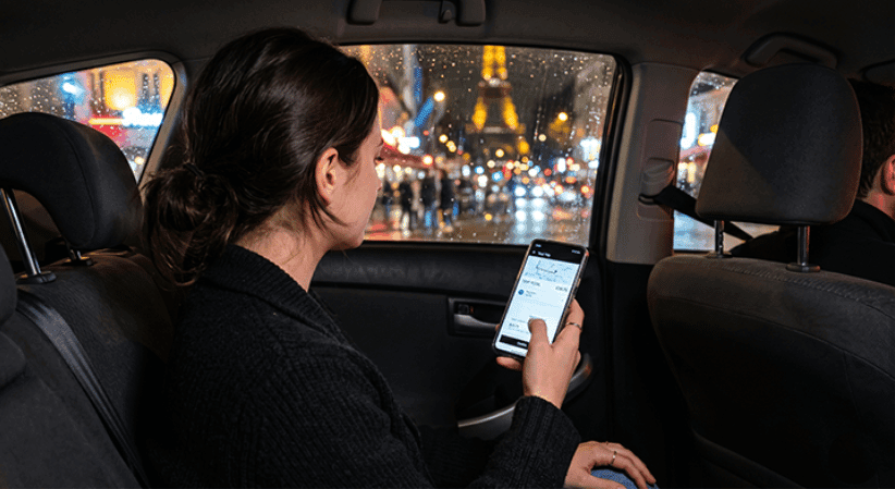 Woman in the back seat of a car at night looking at a rideshare app on her phone, with the Eiffel Tower lit up through the rain-streaked window.
