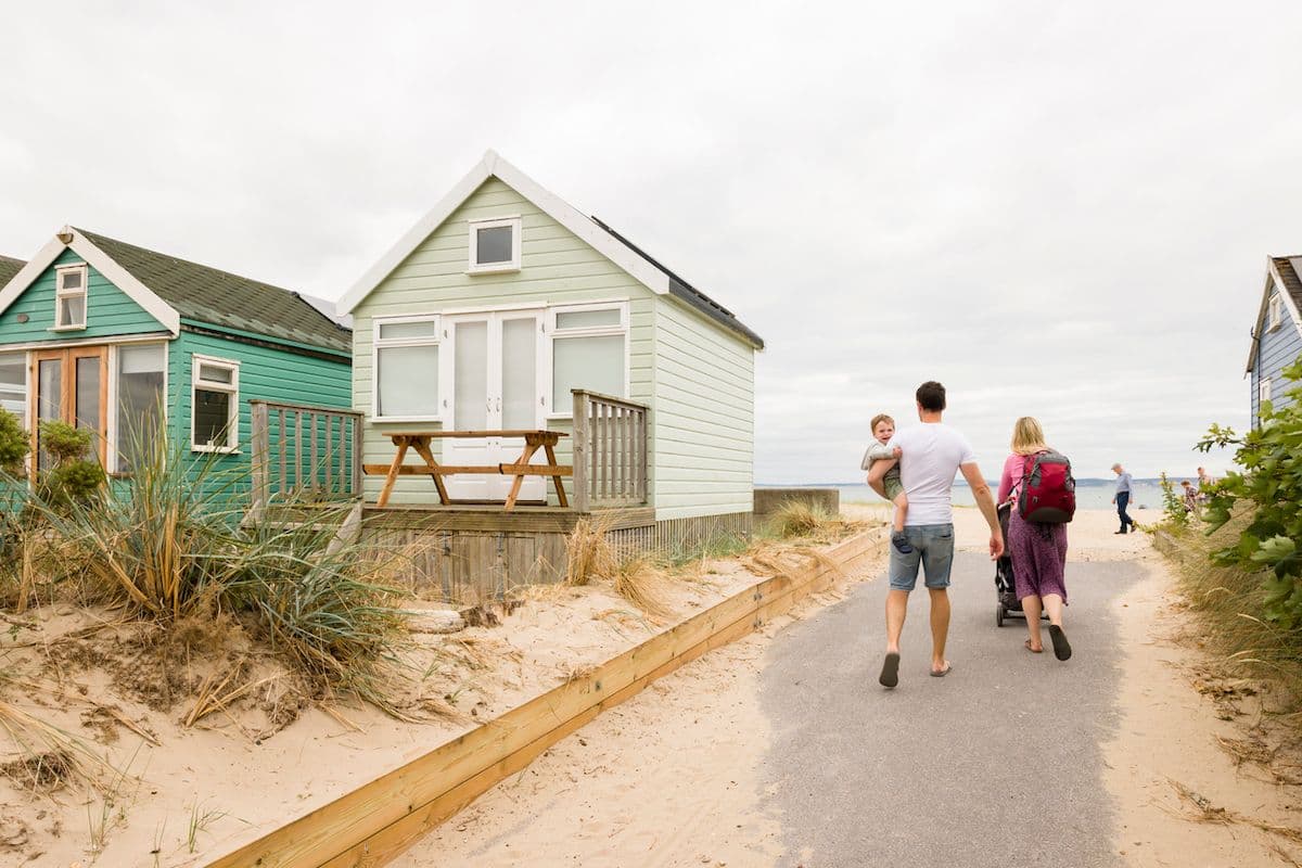 Family walks past pastel beach huts toward the shore on a summer holiday in Dorset, U.K.