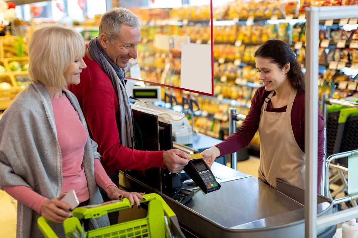 A grocery store checkout scene where a cashier accepts a contactless card payment from an older couple, with shelves of packaged food visible in the background.