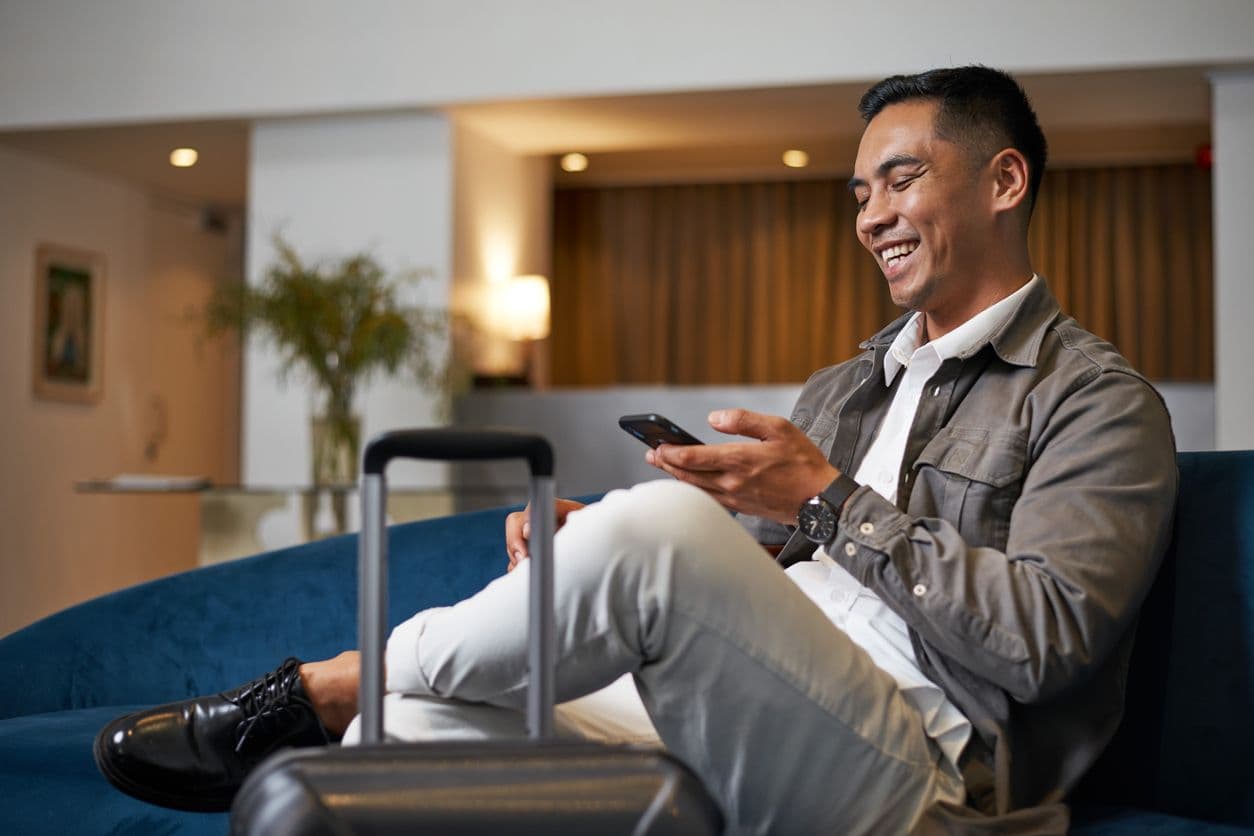 A traveler checks his phone while sitting in an airport lounge with a rolling suitcase.