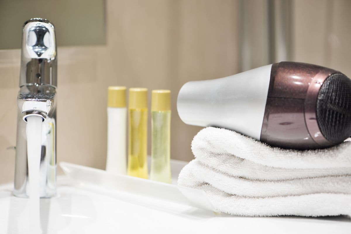 Handheld hair dryer resting on a folded white towel next to mini toiletry bottles and a chrome sink in a hotel bathroom.