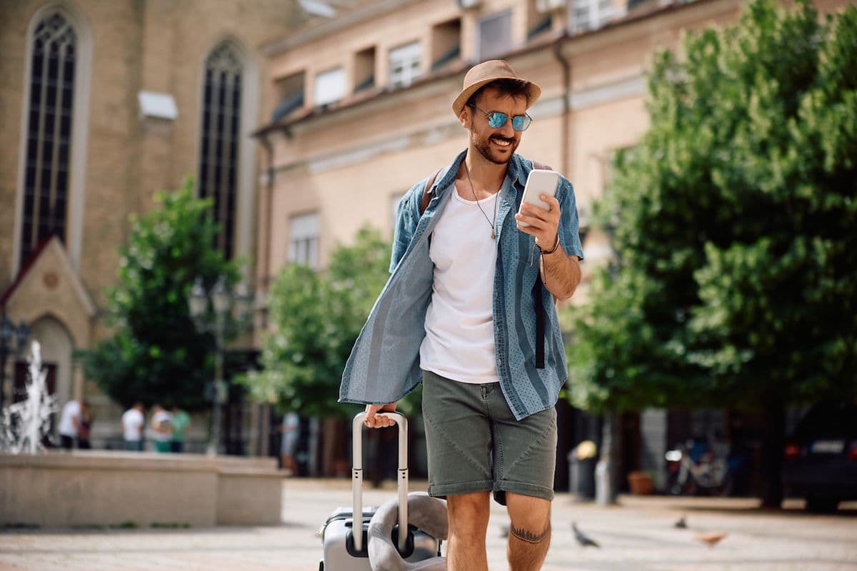 Man walking through a European plaza with a rolling suitcase, smiling at his phone.
