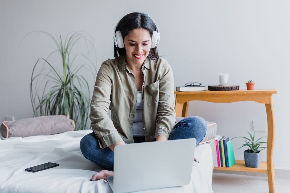 Woman sitting on bed with laptop and headphones, smiling while using a streaming or music service at home.