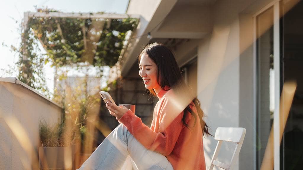 A woman in a coral sweater sits on a sunlit balcony, smiling as she looks at her phone, with potted plants and warm afternoon light around her.