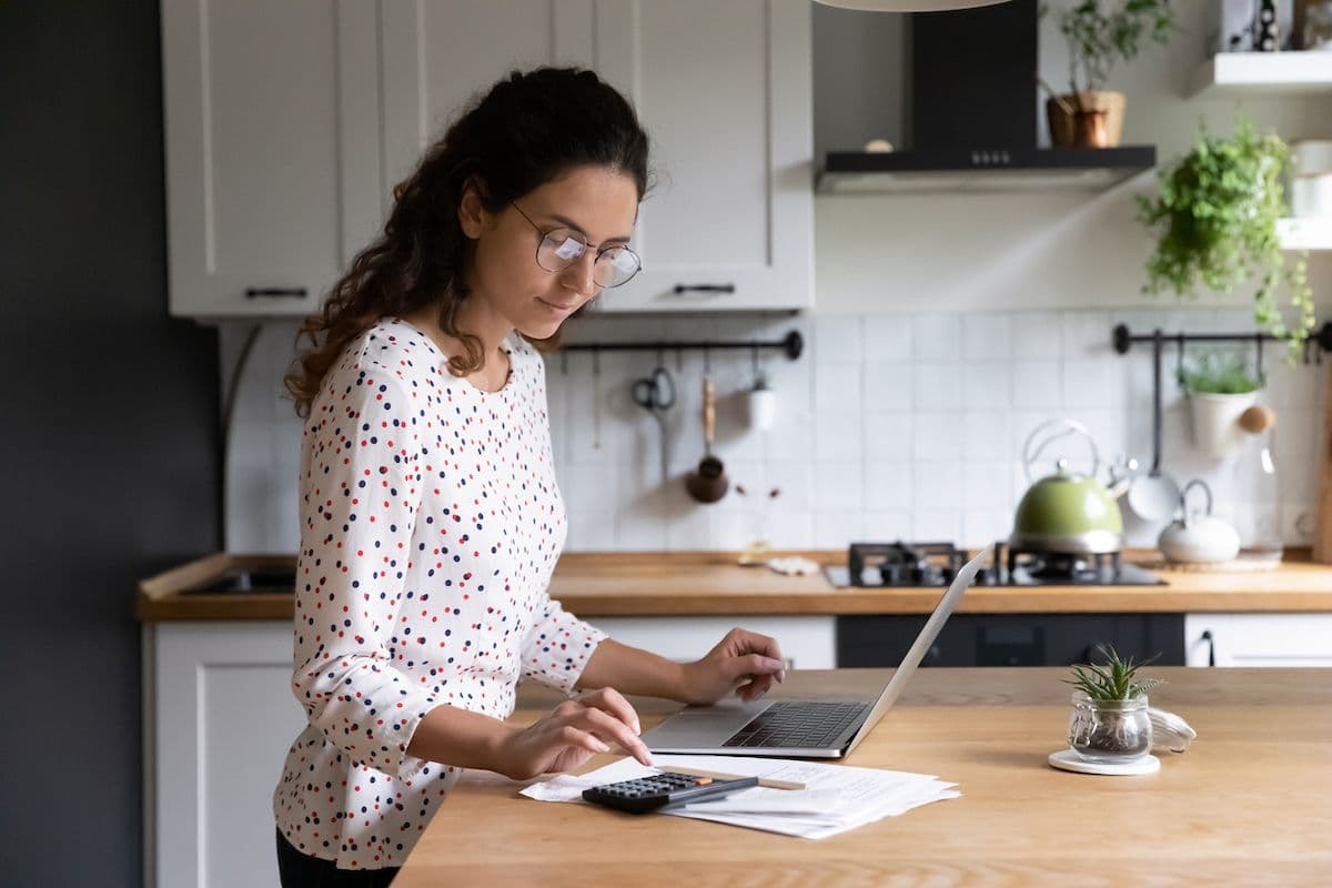 A woman stands at a kitchen counter reviewing documents on a laptop, with a calculator nearby.