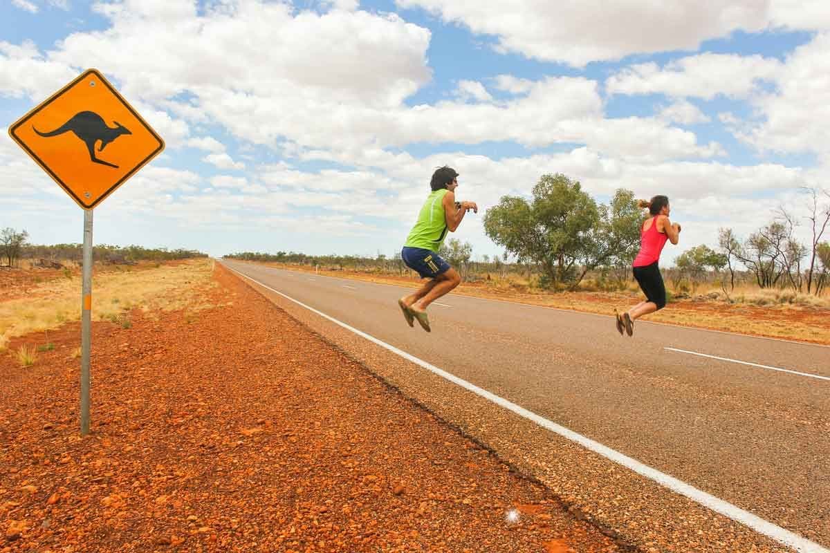Two tourists hop across a road in Australia next to a kangaroo sign.