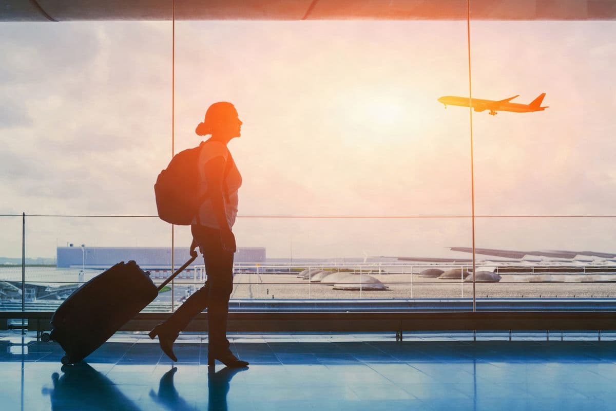 Silhouette of a woman walking through an airport terminal with a rolling suitcase, watching a plane take off through floor-to-ceiling windows at sunset.