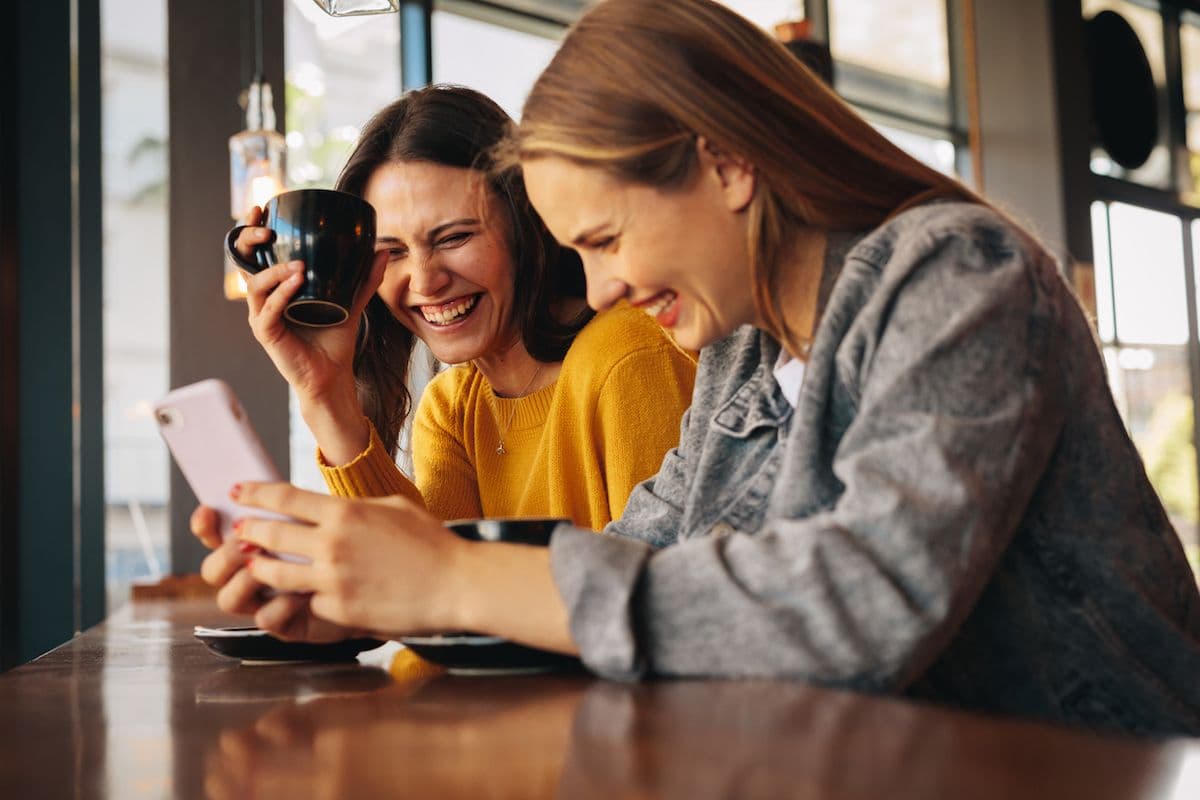 Two friends laughing over a phone at a coffee shop counter