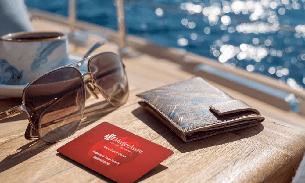 A Medjet Assist membership card beside aviator sunglasses, a coffee cup, and a wallet on a sunny wooden boat deck above blue water
