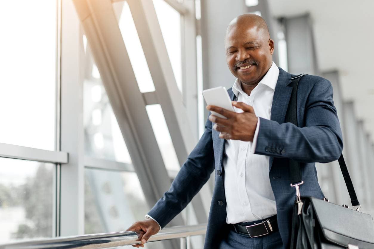 A well-dressed man in a navy suit smiles while looking at his smartphone inside a bright airport terminal, with large windows and a briefcase over his shoulder.