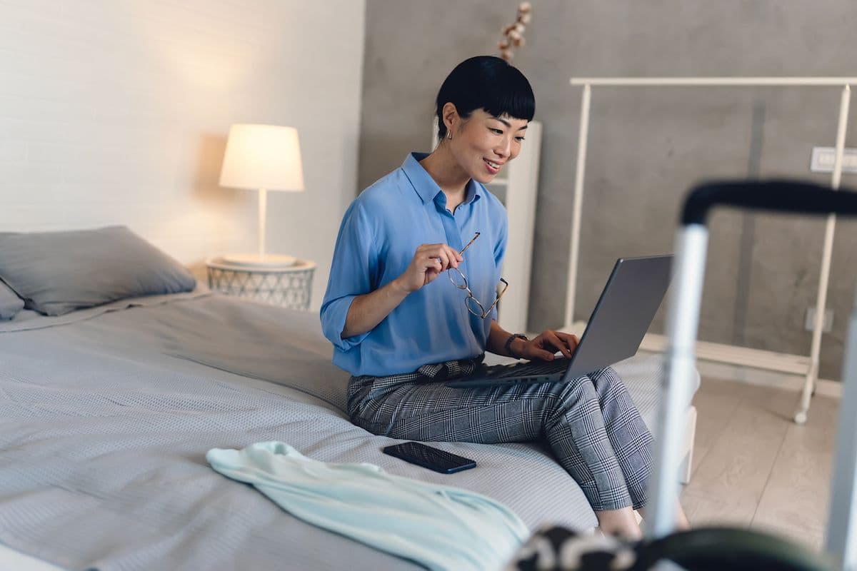A woman sits on a hotel bed with a laptop, holding her glasses, with a suitcase and folded clothing nearby.