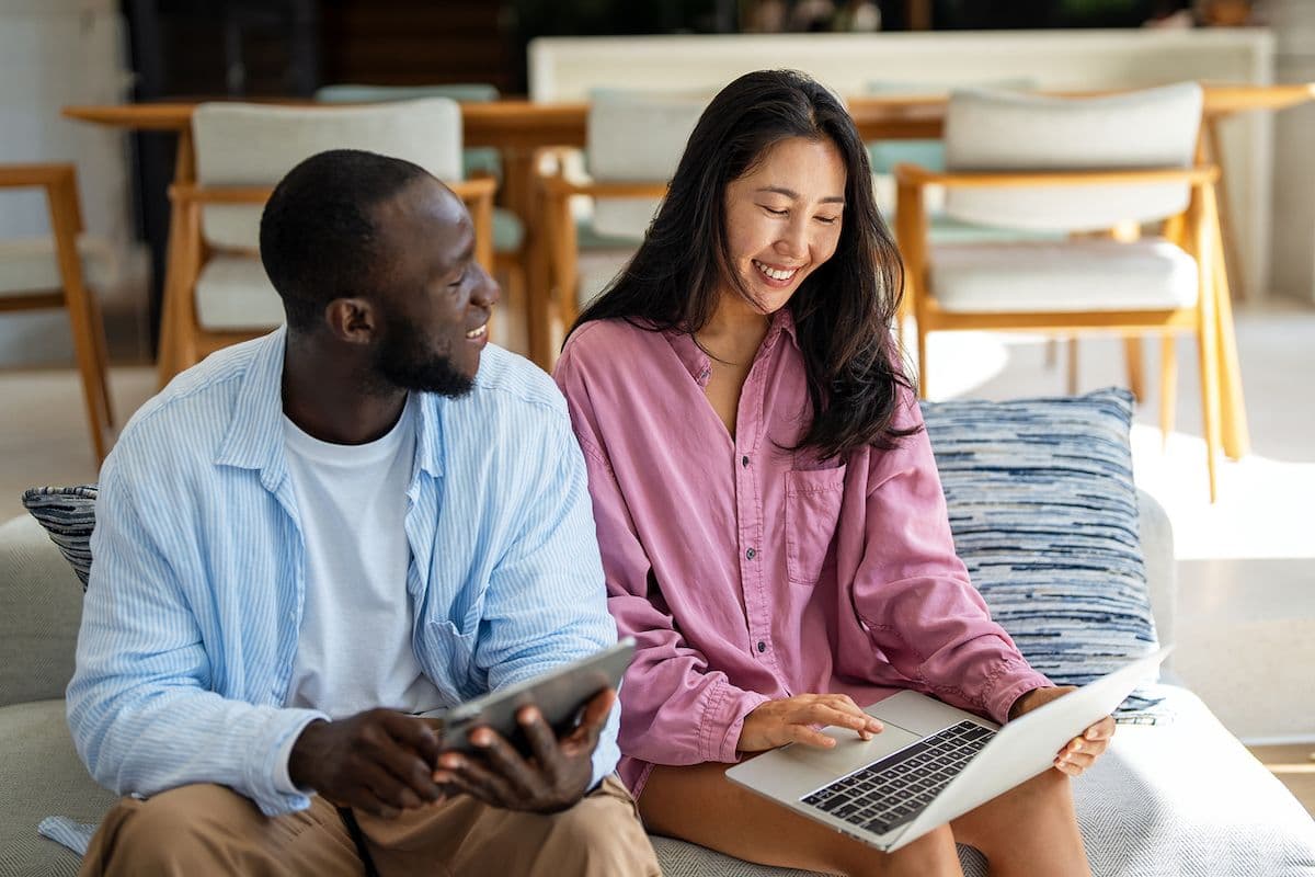 Two adults sitting on a couch compare travel options on a laptop and tablet while planning a trip together.