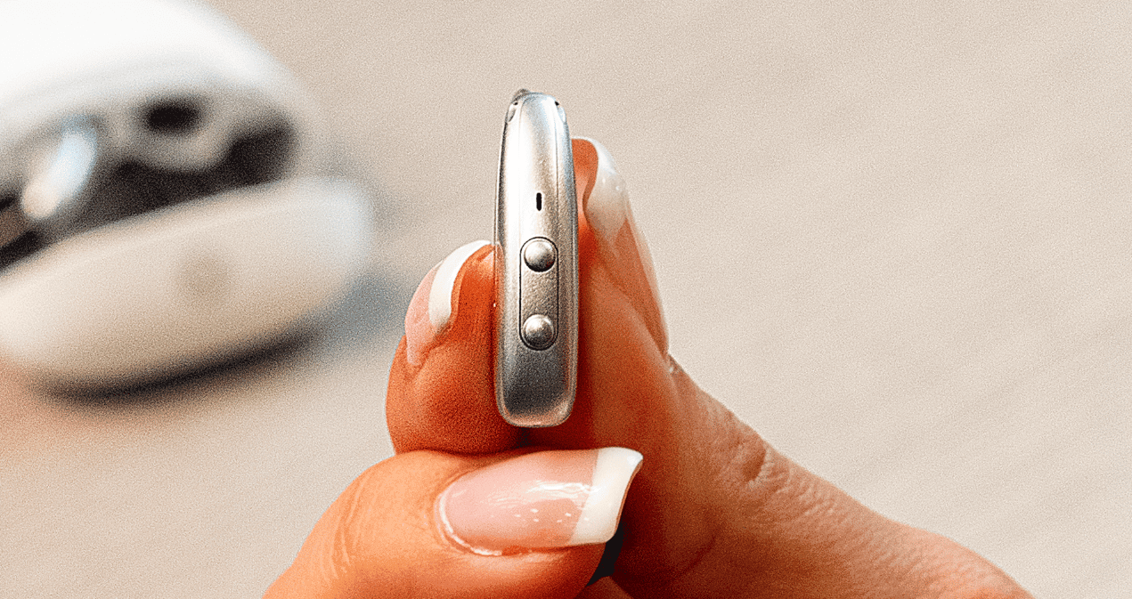 Close-up of a hand holding a small metallic earbud or hearing device, showing side buttons and microphone ports, with another device blurred in the background.