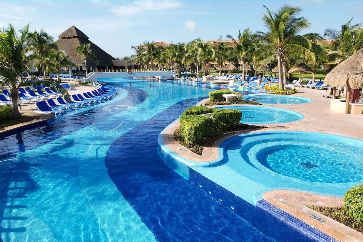 Large resort-style hotel pool with curved turquoise waterways, palm trees, and rows of blue lounge chairs under thatched-roof buildings in a tropical setting