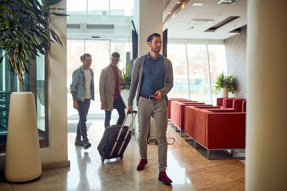 A business traveler wheels a suitcase through a bright, modern hotel lobby while two others follow with luggage
